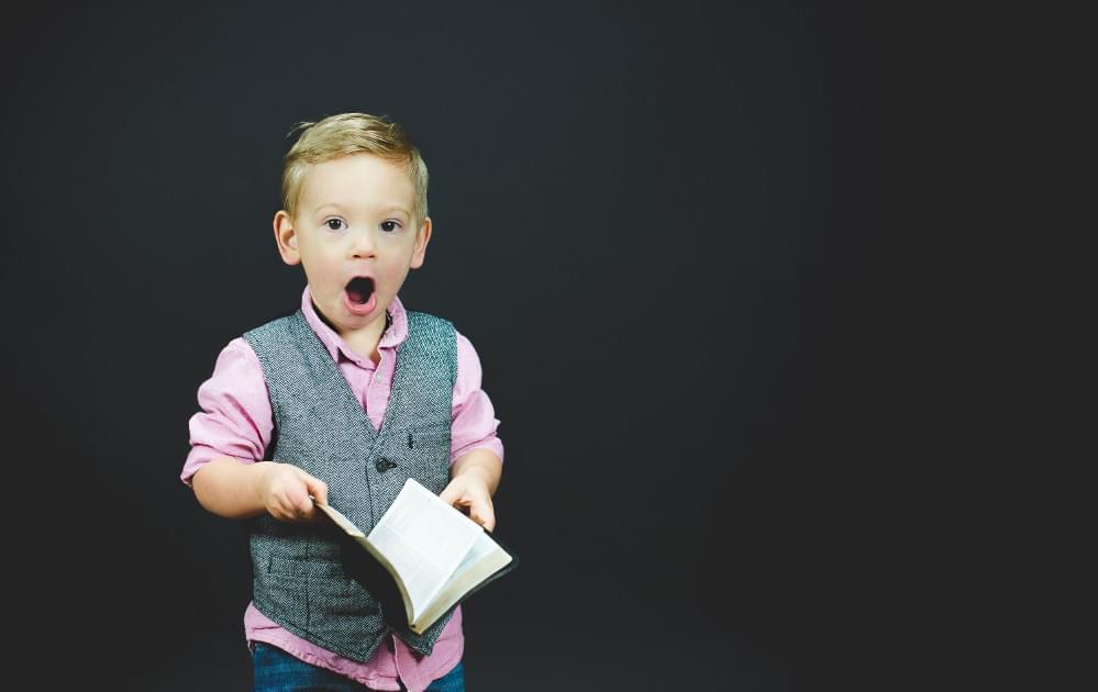 Surprised child wearing gray vest holding a book on black background